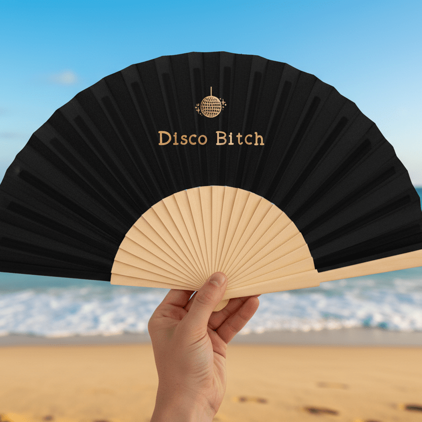 Black fan with 'Disco Bitch' text held by a hand on a beach