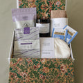Gift box with self-care items including a candle, tea, and skincare products on a floral-patterned surface.