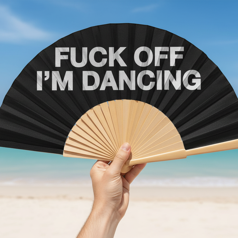 Hand holding a black fan with 'FUCK OFF I'M DANCING' text against a beach background